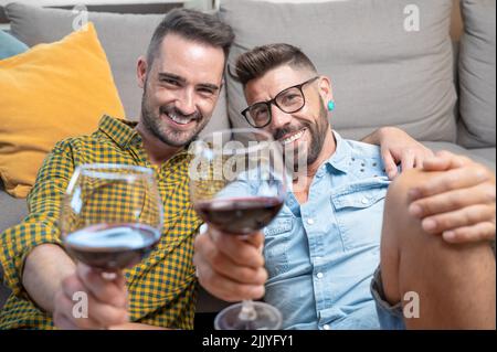 Schwule Paare in der Liebe sitzen zu Hause auf dem Boden und rösten mit Rotwein. Hochwertige Fotografie. Stockfoto