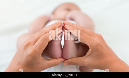 Mama hält einen molligen Fuß des kleinen Babys auf weißem Laken-Hintergrund. Niedliches Neugeborenes im Alter von 3 Monaten. Junge schläft auf dem Bett der Mutter. Familie mater Stockfoto