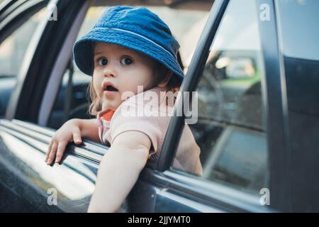 Baby-Mädchen, das ihren Kopf aus einem Autofenster ragt und zurückblickt, während er und seine Familie auf eine Reise gehen. Roadtrip im Sommer. Reisen mit Kindern. Stockfoto