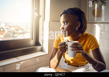 Eine denkende und Kaffee trinkende schwarze Frau genießt ihre morgendliche Routine, während sie an einem hellen sonnigen Tag vor einem Fenster blickt. Schöne weibliche Looks Stockfoto