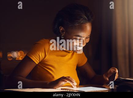 Glückliche, lächelnde Frau, die am späten Abend zu Hause Rechnungen, Darlehen und Online-Shopping auf ihrem Tablet bezahlt. Einzelne afroamerikanische weibliche herunterladen Stockfoto