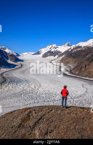 Eine Person in einer roten Jacke steht auf einem großen Felsen mit Blick auf den Lachsgletscher in den Coast Mountains von British Columbia Stockfoto