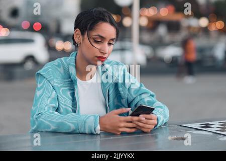 Traurige, depressive und gestresste Frau mit psychischen Problemen, die am Telefon SMS schreibt, während sie im Café im Freien sitzt. Junge Frau bekommt negative Antwort oder Stockfoto