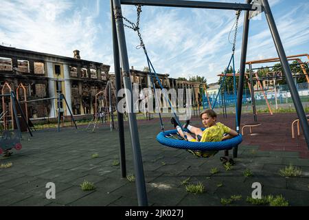 Ein kleiner Junge spielt auf einer Schaukel auf einem Kinderspielplatz neben den Ruinen einer Schule in Charkiw. Charkiw gilt als zweitgrößte Stadt der Ukraine, war eines der wichtigsten strategischen Ziele der russischen Invasion und seit Kriegsbeginn hat die Stadt mehrere Raketenangriffe erlitten, da die Beamten die Stadt als "Heldenstadt der Ukraine" bezeichneten. Stockfoto