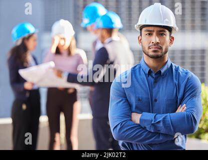 Ein junger Geschäftsmann stand mit gefalteten Armen und trug einen Hardhut, während seine Kollegen hinter ihm standen. Stockfoto