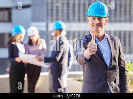 Wir bekamen den Gebäudeteil abgedeckt. Ein reifer Geschäftsmann stand in der Stadt und zeigte einen Daumen nach oben, während er einen Hardhut trug. Stockfoto