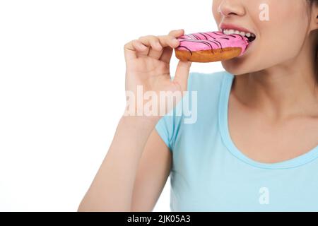 Nicht erkennbare junge Frau trägt ein blaues T-Shirt und beißt appetitlichen Donut, während sie vor weißem Hintergrund steht, Nahaufnahme Stockfoto