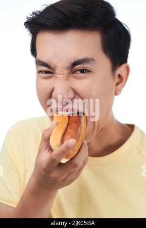 Porträtaufnahme eines jungen asiatischen Mannes, der seine Nase faltet und auf die Kamera schaut, während er Hot Dog isst, isoliert auf weißem Hintergrund Stockfoto