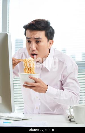 Schockiert asiatischen weißen Kragen Arbeiter Blick auf den Computer-Bildschirm, während Instant Nudeln mit Essstäbchen essen, Taille-up-Porträt aufgenommen Stockfoto