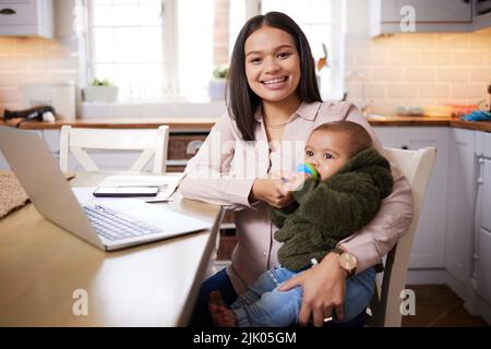 Multitasking von seiner besten Seite. Eine junge Frau, die einen Laptop benutzt, während sie sich zu Hause um ihr entzückendes Baby kümmert. Stockfoto