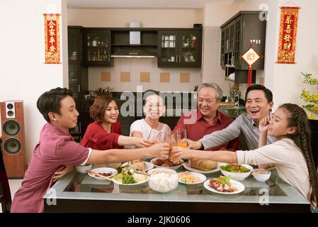 Fröhliche vietnamesische Familie, die mit Gläsern Saft toaste. Schriftrollen, die Glück und Reichtum im Hintergrund wünschen Stockfoto