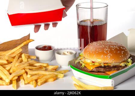 Frische leckere Burger, Cola und pommes auf weißem Hintergrund. Die Hand einer Frau gibt eine Kiste mit Lebensmitteln, Seitenansicht. Zwei verschiedene Fleisch-Burger in einer Schachtel mit Stockfoto