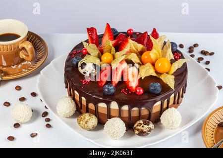 Schokoladenkuchen mit Erdbeeren, Moor-Wortleberry, Physalis, weißen Candiesand, Osterquai-Eier auf weißem Hintergrund mit einer Tasse duftenden Kaffee für E Stockfoto