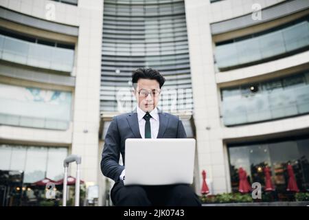 Vietnamesischer junger Geschäftsmann, der vor dem Bürogebäude sitzt und an einem Laptop arbeitet Stockfoto