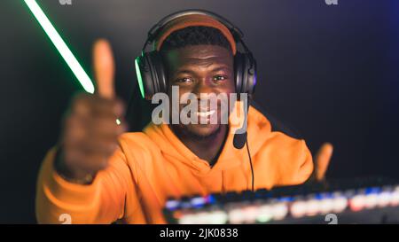 Junger schwarzer Mann mit Mütze und Headset, der lächelnd in die Kamera schaute und die farbenfrohe Tastatur hochhielt und den Daumen hoch gab. Hochwertige Fotos Stockfoto