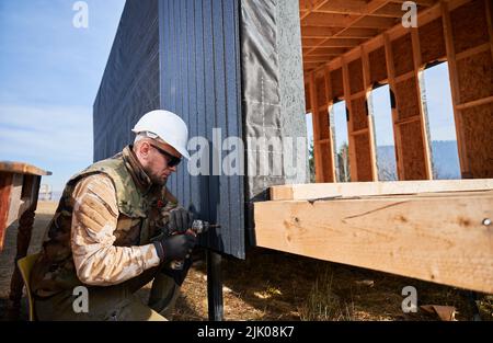 Männlicher Baumeister, der schwarze Wellblech installiert, das als Fassade des zukünftigen Häuschens verwendet wird. Nahaufnahme eines Arbeiters, der ein Holzhaus baute. Zimmerei- und Konstruktionskonzept. Stockfoto