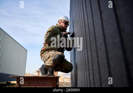 Männlicher Baumeister, der schwarze Wellblech installiert, das als Fassade des zukünftigen Häuschens verwendet wird. Mann Arbeiter Gebäude Holzrahmen Haus. Zimmerei- und Konstruktionskonzept. Stockfoto