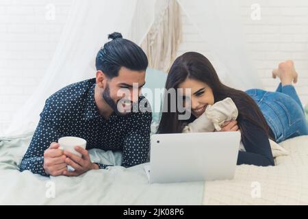 Ein Paar liegt im weißen Schlafzimmer und entspannt sich gemeinsam vor einem Laptop und trinkt Kaffee. Innenaufnahme. Hochwertige Fotos Stockfoto