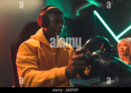 Schwarzer Teenager, der in einem Spielstuhl sitzt und Kopfhörer trägt und Rennspiele mit dem Lenkrad spielt. Dunkelrote und grüne Beleuchtung. Hochwertige Fotos Stockfoto