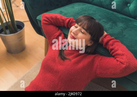 Ruhige glückliche junge Brünette Frau trägt einen roten Pullover und entspannt sich auf einer Couch mit geschlossenen Augen. Hochwertige Fotos Stockfoto