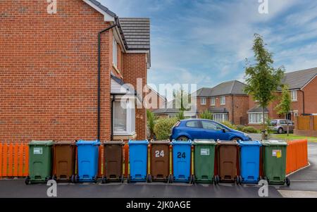 Eine Reihe farbiger Mülltonnen zum Recycling von Kunststoff-, Papier- und Gartenabfällen, neben einem orangefarbenen Zaun und vor einem modernen Haus. Stockfoto