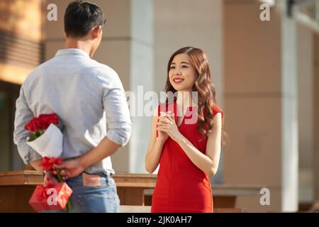 Hübsche asiatische Frau trägt rotes Kleid im Freien stehen und erwarten wunderbare Überraschung, während ihr Freund versteckt Strauß von roten Rosen und Geschenkbox hinter seinem Rücken Stockfoto