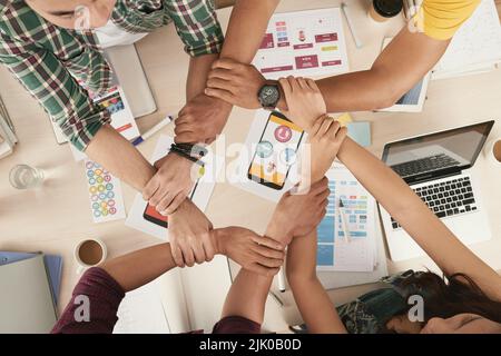 Junge Menschen halten sich gegenseitig die Hände in einer Kette über dem Tisch mit Dokumenten für mobile Apps Stockfoto