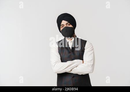 Südasiatischer Mann mit Turban und Gesichtsmaske, der die Kamera isoliert über der weißen Wand ansieht Stockfoto