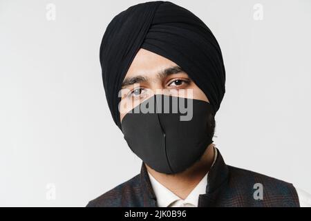 Südasiatischer Mann mit Turban und Gesichtsmaske, der die Kamera isoliert über der weißen Wand ansieht Stockfoto