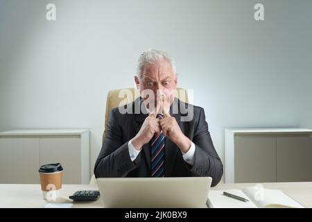 Nervöser, reifer Unternehmer, der in klassischem Anzug auf dem Laptop-Bildschirm mit Konzentration sitzt, während er am Schreibtisch sitzt, Porträtaufnahme Stockfoto