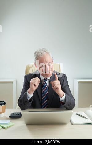 Fröhlicher, reifer Manager, der mit einem breiten Lächeln und einem freundlichen Fingerschütteln auf den Computerbildschirm blickt, während er einen Videoanruf an einen Kollegen führt, und die Inneneinrichtung eines modernen Büros im Hintergrund Stockfoto