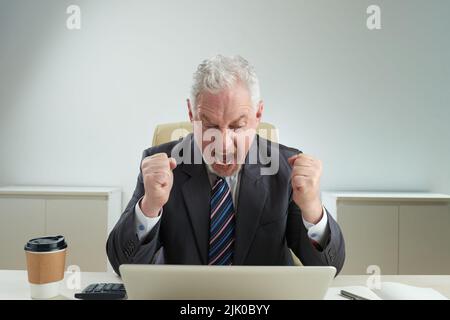 Verärgerter Geschäftsmann im klassischen Anzug, der die Hände in die Fäuste hält und nach einem Scheitern schreit, die Inneneinrichtung des modernen Büros im Hintergrund Stockfoto