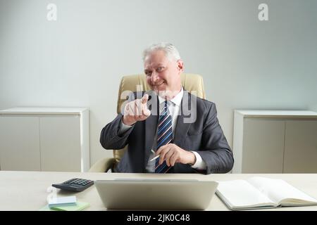 Ein gutaussehender Senior Manager mit einem dutzigen Lächeln, der auf die Kamera zeigt, während er am Schreibtisch sitzt und an einem vielversprechenden Projekt arbeitet, einem Porträt mit Hüfthöhe Stockfoto