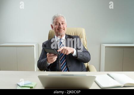 Ein gutaussehender, älterer Finanzmanager, der die Kamera mit einem dutzenden Lächeln anschaut, während er den Rechner benutzt, und das Innere des modernen Büros im Hintergrund Stockfoto