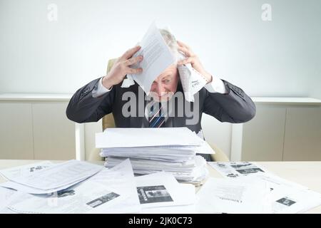 Verärgerter Geschäftsmann, der seinen Kopf mit Dokumenten bedeckt, während er von der Arbeit überwältigt am Schreibtisch sitzt, Porträtaufnahme Stockfoto