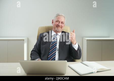 Porträt eines fröhlichen älteren Geschäftsmanns, der am Schreibtisch sitzt und den Daumen hochzeigt, auf dem Hintergrund der Büroeinrichtung Stockfoto