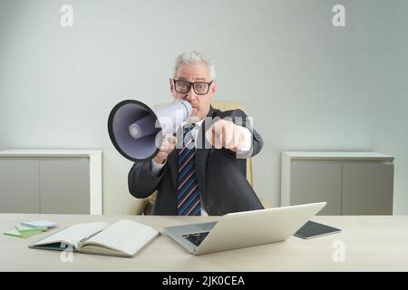 Bärtiger Unternehmer, der einen Anzug mit Megaphon trägt und auf die Kamera zeigt, während er am Schreibtisch eines modernen Büros sitzt und ein Porträt mit Hüfthöhe zeigt Stockfoto