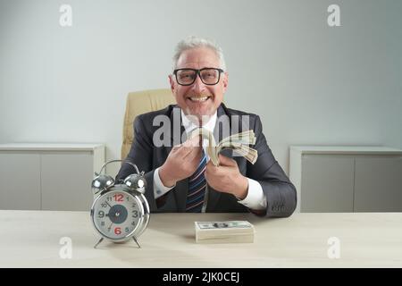 Fröhlicher Unternehmer in klassischem Anzug und Brillen, der mit einem Lächeln auf die Kamera blickt, während er ein Geldscheinbündel in der Hand hält und auf dem Schreibtisch einen Wecker stellt Stockfoto