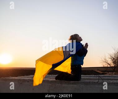 Silhouette eines Jungen auf den Knien, hinter dem Rücken des Kindes flattert die Flagge der Ukraine im Wind. Hände gefaltet im Gebet, appellieren an Gott. Kinder gegen Krieg. Ukr Stockfoto