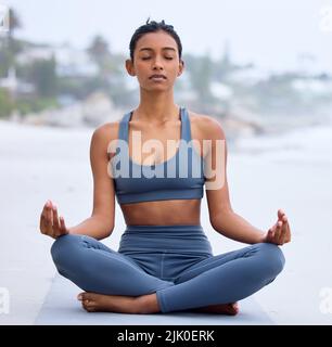 Innerer Frieden ist der Schlüssel. Ganzkörperaufnahme einer attraktiven jungen Frau, die am Strand beim Yoga-Üben meditiert. Stockfoto