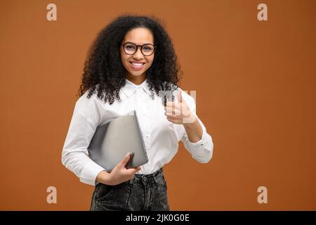 Junge afroamerikanische Frau mit Afro-Frisur in eleganter Freizeitkleidung, isoliert auf Braun stehend und mit großem Finger, Laptop-Computer tragend, lächelnd, weibliche Büroangestellte, Studentin Stockfoto