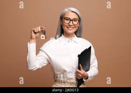 Portrait of adorable Asian Woman Holding a car key, presenting smart phone isoliert auf braunem Hintergrund in Studio mit Copy Space Stockfoto