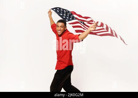 Fröhlicher afroamerikanischer Typ, der in Händen amerikanische Flagge hält und isoliert auf weißem Hintergrund läuft, multirassischer ausländischer Student mit US-Flagge, Unabhängigkeitstag, Feiertag Stockfoto