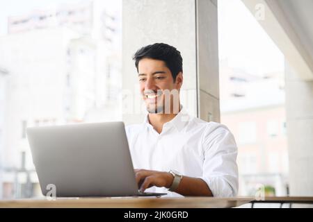 Ein lateinischer Entwickler, Freiberufler und Büroangestellter verwendet einen Laptop, der draußen am Schreibtisch sitzt. Ein junger, multirassischer Kerl tippt auf der Tastatur, antwortet auf E-Mails und kommuniziert mit dem Kunden Stockfoto