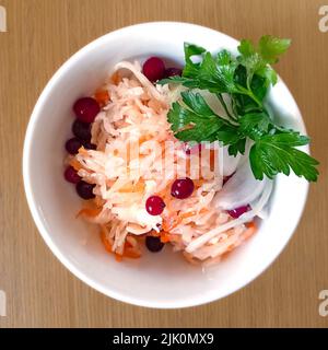 Sauerkrautsalat mit Preiselbeeren und Karotten mit einem Zweig grüner Petersilie auf einem weißen Teller auf dem Tisch. Die Idee einer gesunden Ernährung. Fermentierte Lebensmittel Stockfoto