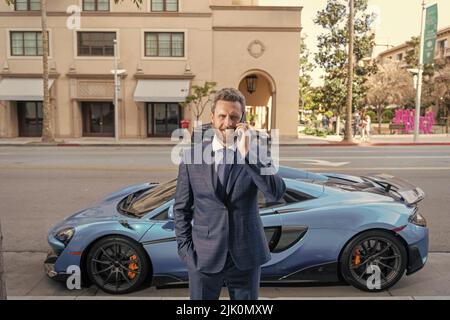 Selbstbewusster Händler spricht auf dem Smartphone neben dem Auto Stockfoto