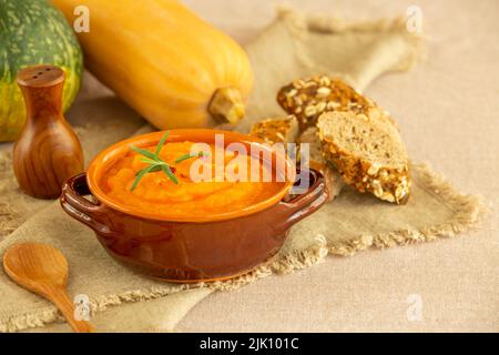 Kürbis- und Karottensuppe in Tonschüsseln und Bio-Kürbisse mit schwarzem Brot auf Leinenhintergrund. Vegetarische Herbstsahnesuppe aus Kürbis mit Samen. Stockfoto