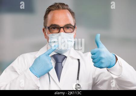 Der Arzt trägt eine medizinische Gesichtsmaske und medizinische Handschuhe und sticht zu seiner hygienischen Ausrüstung und zeigt den Daumen nach oben Stockfoto