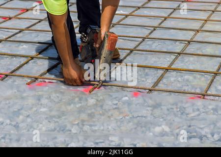 Der Bauarbeiter verdreht die Drahtverstärkung mit einem Bewehrungswerkzeug in Stahlstäbe, um aus Stahlstäben ein Zementfundament zu bilden. Stockfoto