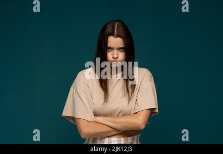 Porträt eines verärgert jungen schönen Mädchen in einem beigen T-Shirt stehen mit geschwollenen Wangen und Arme isoliert auf blauem Hintergrund gefaltet. Emotionen der Wut Stockfoto